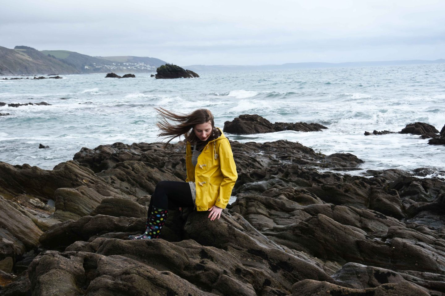 melissa carne in seasalt yellow coat sitting on rocks with windy hair in cornwall