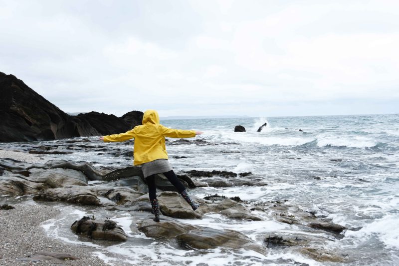 melissa carne balancing on rock in yellow coat by the sea in cornwall